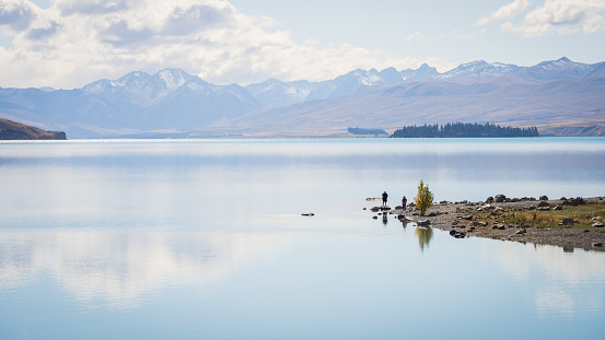 Wide alpine scenery with people walking on the bank of a lake with mountains in backdrop,New Zealand.
