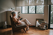 sad asian senior woman sitting in living room looking outside window with hot drink