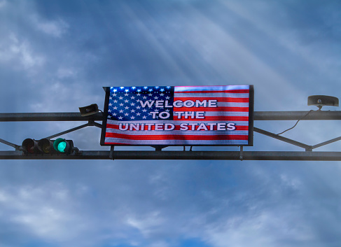 Digital Sign with American Flag stating Welcome To The United States against a cloudy sky.The image was taken at the Otay Mesa Border crossing betwen the USA and Mexico