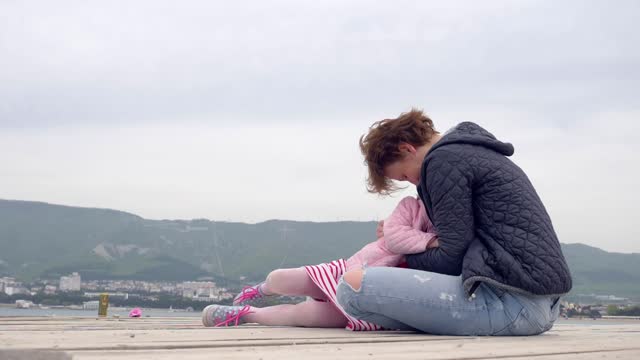 On a cloudy seaside pier, a mother and daughter share an embrace, enveloped in a heartwarming talk