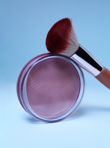 Blush and blush brush on light gray background. Pink blush in compact package with transparent lid. The brush has brown handle and synthetic bristles.