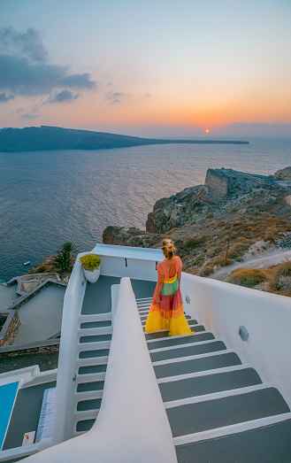 Young tourist woman enjoys the Caldera and Aegean Seaview and the holiday as she descends the traditional white and grey stairs to the ocean looking sun setting in Oia or Ia on a summer time in Santorini, Greek Islands, Cyclades, Greece.