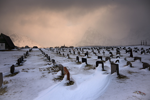 A grave yarad in winter with tomb stones buried in snow and a church at the background under mysterous cloudy sky