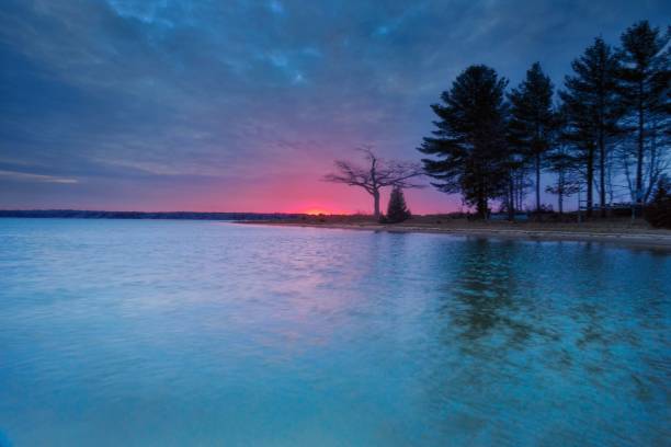Higgins Lake, Michigan. 4/9/24. A tranquil lake reflects the vibrant hues of sunset, with broad streaks of pink and orange light breaking through the cloud cover. A tree-lined shoreline gently meanders into the distance on the right, providing a stark contrast to the open waters. Drone point of view.