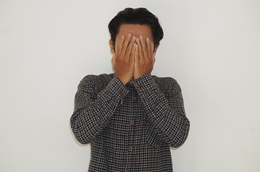 Young asian man in black shirt covering his face with hands on white background.