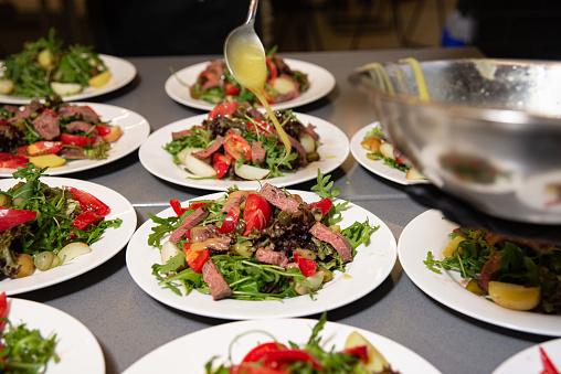 Chef precisely drizzling dressing over steak salads, beautifully arranged on plates in a busy commercial kitchen setting.