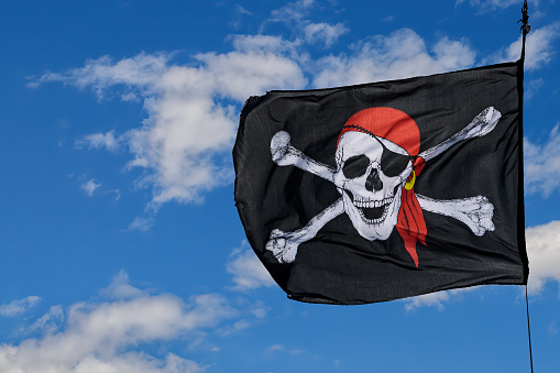 A black, white, and red generic Jolly Roger flaps in the winds against a partially cloudy blue sky. The skull has one eye patch and one gold earring.