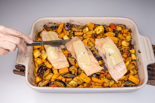 A baking dish with oven roasted vegetables and salmon steaks with melted parmesan cheese. and a hand with a knife checks if cooked.