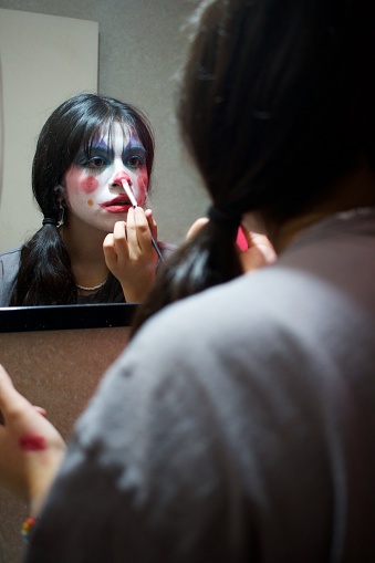 clown girl making up her face in the mirror of a dark bathroom, side view