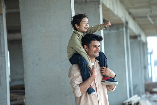 Smiling young father carrying cheerful son on shoulders and looking away while standing at construction site