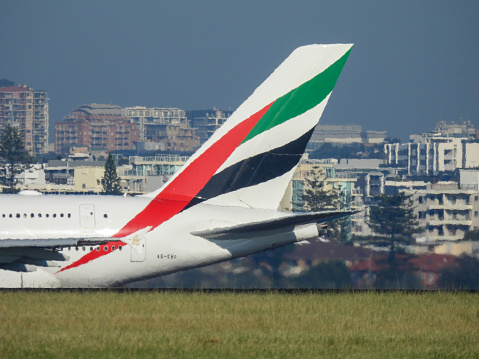 The vertical stabiliser of an Emirates Airbus A380-842 plane, registration A6-EVD, taxiing in preparation for departure from Sydney Kingsford-Smith Airport as flight EK412 to Christchurch.  In the background are residential buildings. This image was taken from Mill Stream Lookout, Botany Bay on a sunny morning on 30 March 2024.