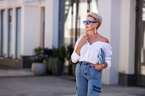 Beautiful blonde fashion mature woman outdoors in city street wearing trendy white shirt blouse, cargo jeans pants and fashionable accessories, stylish sunglasses and clutch. Street style portrait.