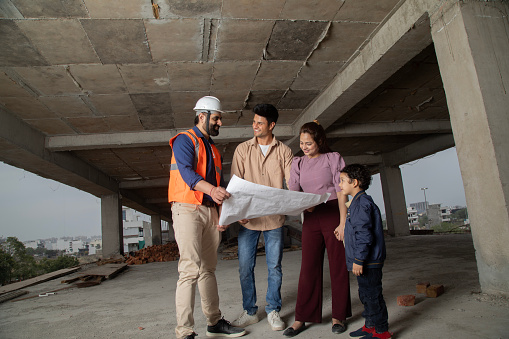 Male architect showing and discussing new apartment blueprints to young parents and son at construction site