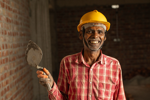Portrait of smiling senior male mason with arms crossed holding trowel and standing by brick wall at construction site