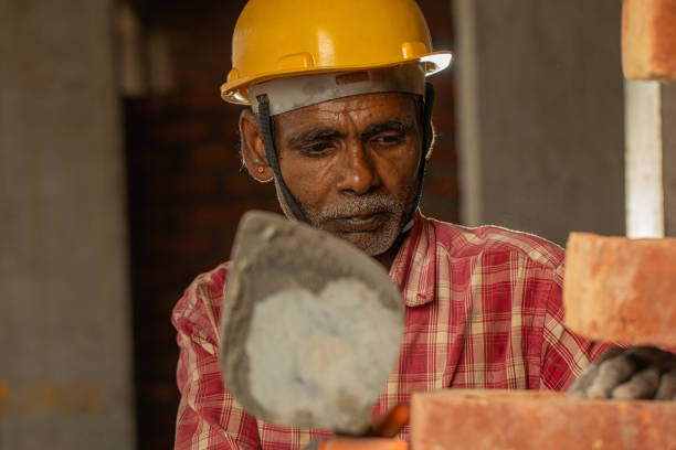 Portrait of senior male mason in hardhat laying bricks with trowel on exterior walls at construction site