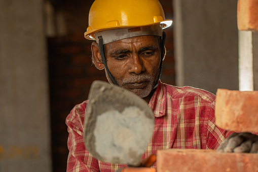 Portrait of senior male mason in hardhat laying bricks with trowel on exterior walls at construction site