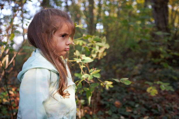 A young girl looks upwards with an expression of frustration and annoyance, her arms crossed tightly against her chest. The natural light captures her features against a dark, leafy background, emphasizing the depth of her irritation. Her slightly parted lips and furrowed brow convey a strong sense of being fed up, as if she's reached the limit of her patience. This image powerfully portrays the raw emotions of exasperation and discontent in a candid outdoor setting.