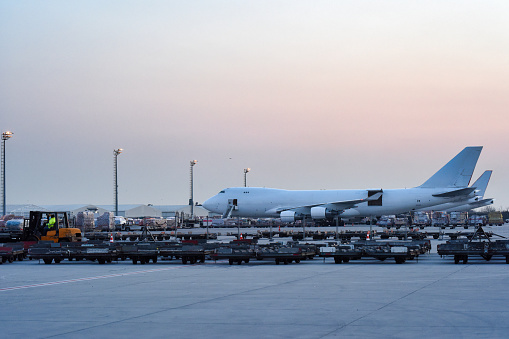 A large cargo airplane sits on the tarmac amid the hustle of baggage and cargo vehicles preparing for the next flight, under the soft glow of dawn.