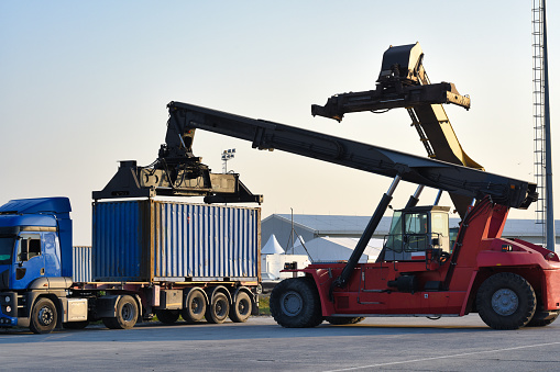 A red reach stacker lifting a blue shipping container onto a truck trailer under the clear daylight sky at a cargo handling facility.