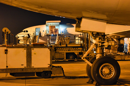 Workers busy loading cargo onto an aircraft at night, with the foreground showcasing ground support equipment and part of the plane's undercarriage.