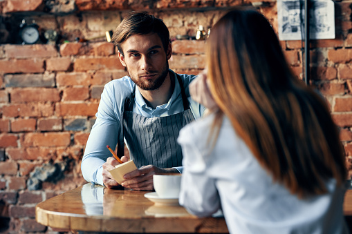 a man with a cup of coffee and all aprons at a cafe table and a woman in a shirt and red skirt cropped view. High quality photo