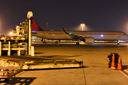 An airplane stands by for its journey on the lit apron of an airport, with ground support equipment and safety cones in the foreground, capturing the essence of nighttime airport operations.