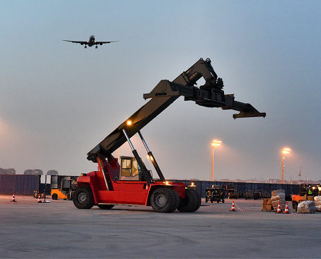 A reach stacker stands poised on the tarmac as an airplane approaches landing in the background, depicting a moment of ground-to-air transition at an airport.