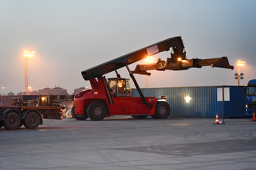 A reach stacker lifts cargo at twilight, with the lights of the terminal shimmering in the background, highlighting the ongoing activity in the freight yard.