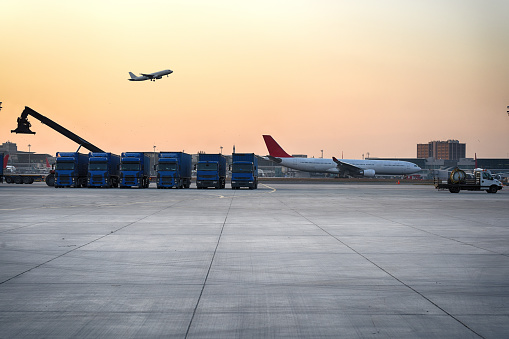 Trucks aligned at an airport cargo area with a reach stacker in the foreground and an airplane taking off into the dusk sky in the background.