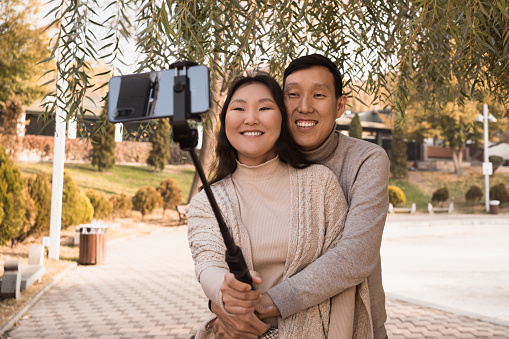 Happy smiling hugging young Asian couple in love taking selfie photo by phone under tree in autumn city park