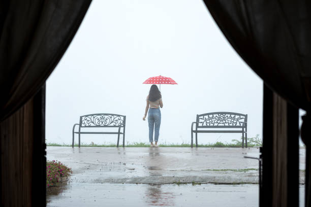 Viewed through a curtain's opening, a lone individual stands with a red polka-dotted umbrella, gazing into the fog-covered landscape, imparting a sense of contemplation and solitude