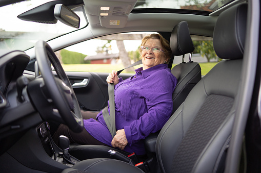 A smiling senior woman, wearing a purple shirt, buckles her seat belt in the car with calmness and confidence