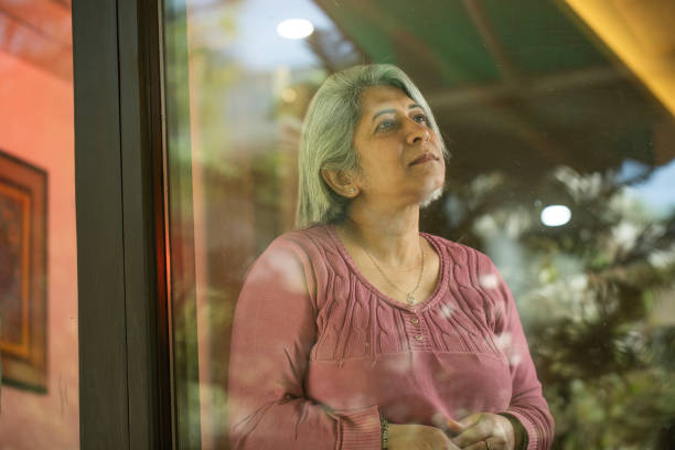 istock Thoughtful senior woman looking through window while standing at house 2127429841