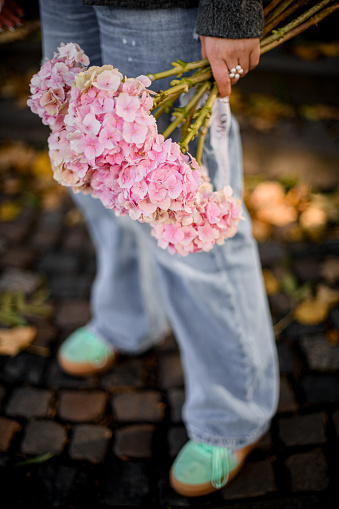 Focus on a bouquet of large-leaved hydrangeas in the hands of a young woman with a flower-shaped ring on her finger on a blurred background outdoors, close-up