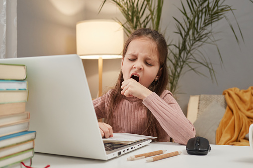 Kid-friendly online study. Digital education at home. E-learning for children. Sleepy tired little girl using laptop and yawning while sitting at table at home