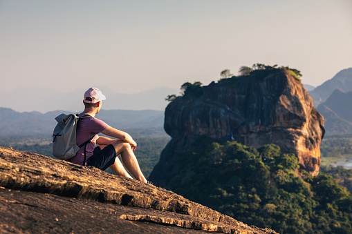 Man with backpack sitting on rock and looking at landscape. Scenery with Sigiriya rock in Sri Lanka.