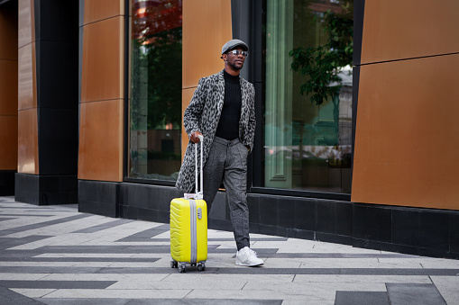 Stylishly dressed male model walks down city street with yellow suitcase in hand. Black man tourist outdoors near hotel building. African american man in trendy coat, cap and Luggage.