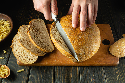 A cook cuts fresh bread with a knife on a kitchen board close-up. Slicing rye bread on the kitchen table or concept of healthy eating and traditional baking.