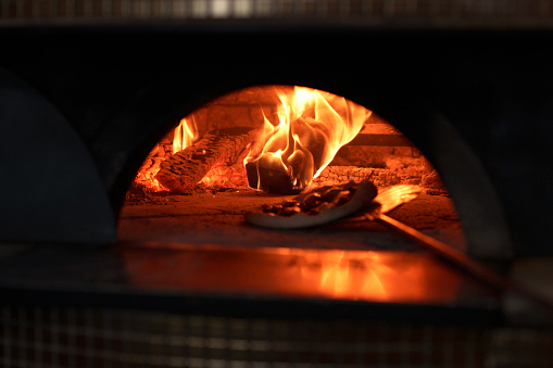 The chef adjusts the pizza paddle inside a brick oven fire. This action ensures that the pizza cooks evenly and achieves the desired level of crispiness and flavor from the intense heat of the oven.