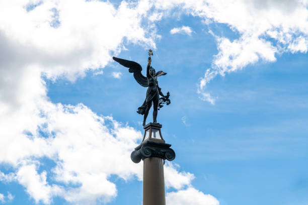 Elevated bronze angel statue with wings spread wide, standing against a backdrop of blue sky and fluffy clouds
