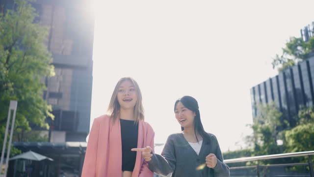 Front view of Happy asian businesswoman and female friend walk beside office in capital city with lights shining behind them.
