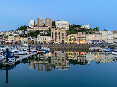 View across Torquay harbour in the centre of Torquay, UK.  Boats can be seen moored in the harbour. People can be seen on the promenade