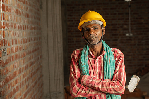 Portrait of senior male mason with arms crossed holding trowel and standing by brick wall at construction site