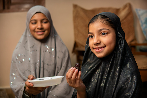 Two happy Islamic girls in hijab eating traditional dates fruits during Ramadan fasting month at home.