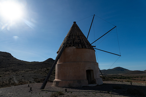 Cabo de Gata, Almeria - Spain - 01-23-2024: An old windmill stands under the blue sky backlit by the sun in the arid landscape of Cabo de Gata, Spain