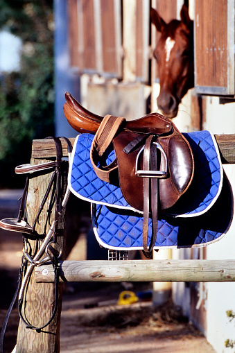 The saddle is brown on the fence in a shallow depth of field. leather saddle, harness for horses. Western saddles for horses on the rack, ready for dressage training. Equestrian sport background.