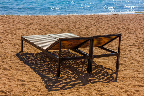 Two empty outdoor lounge chairs on sandy beach near blue water at sunny off-season day.