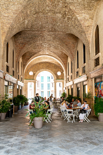 Vicenza, Venetien - Italy - 06-12-2021: Diners enjoy outdoor seating on white chairs between plants under the scenic vaulted arches of the historic architecture of Palladian Basilica