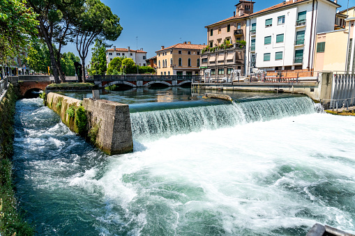 Treviso, Venetien - Italy - 06-13-2021: Vivid view of a waterfall of a weir with rushing water and a fish ladder on the side in Treviso with historic buildings in the background