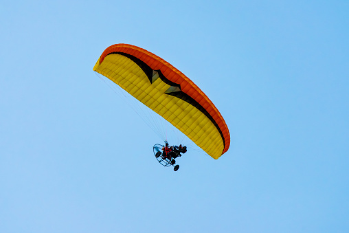 Caorle, Venetien - Italy - 06-14-2021: An orange, yellow, and black paramotor flies high, offering a thrilling view against the clear blue sky